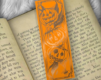 Puede incluir: Marcapáginas naranja con una ilustración en negro y plata de dos calabazas con la palabra "MAD" en el centro. El marcapáginas tiene un borde negro y un diseño dorado.