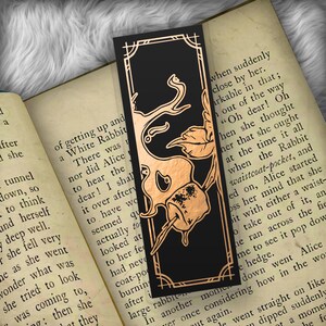 May include: Black bookmark with a copper-colored illustration of a rabbit, a leaf, and a marshmallow. The bookmark has a decorative border and is set against the pages of a book.