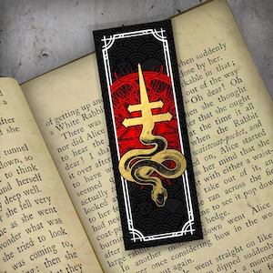May include: A black bookmark with a gold and red design featuring a cross, snake, and other symbols. The bookmark is rectangular with a white border and is placed on an open book.