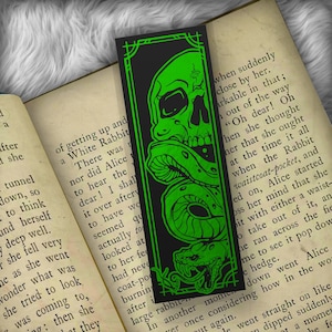 May include: Black bookmark with a neon green skull and snake design. The bookmark has a rectangular shape with a decorative border. The skull is at the top, and the snake winds down the bookmark. The bookmark is placed on top of an open book.