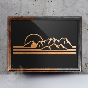 May include: A framed print with a black background and a gold silhouette of mountains and a sun. The mountains are in the foreground and the sun is in the background. The print has a gold and black frame.