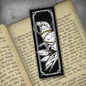 May include: A black and white bookmark featuring a raven with a yellow rope tied around its neck. The raven is perched on a branch with a white background. The bookmark has a black border with a decorative pattern.