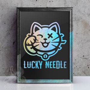 May include: Framed art print featuring a stylized cat design with a winking eye and cross-shaped detail. The cat is in a rainbow holographic color, set against a black background. The text "LUCKY NEEDLE" is displayed below the cat.