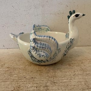 May include: A ceramic bowl shaped like a swan with a white base and blue painted feather details. The swan head extends upward from one side, while the wings are sculpted along the sides of the bowl. The interior is smooth and white.