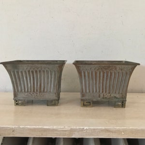 Puede incluir: Dos macetas cuadradas de metal antiguas con un acabado desgastado y envejecido. Las macetas tienen un diseño decorativo acanalado y se apoyan en cuatro pequeños pies decorativos.