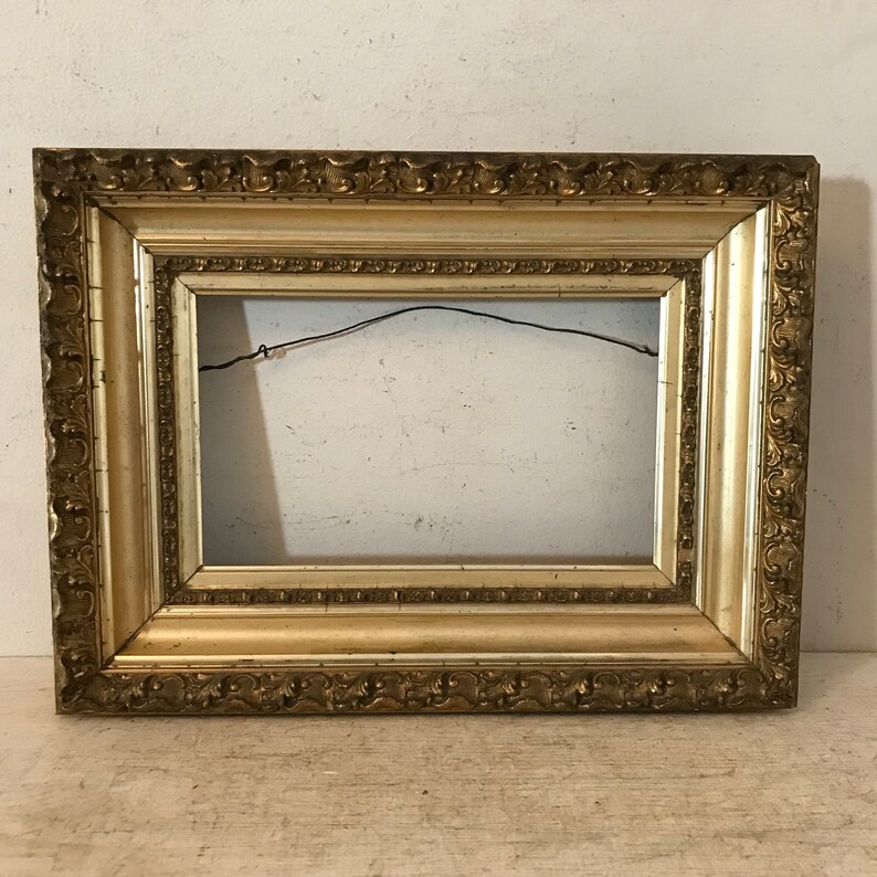 May include: A gold-colored picture frame with an ornate design. The frame has a rectangular shape and a slightly raised edge.