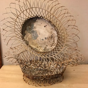 May include: An antique, white wire basket with a decorative, sunflower-like design. The basket has a round, metal insert with peeling paint. The basket is likely vintage and shows signs of age and wear.