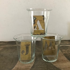 May include: Three clear glass cups with brass stencils inside. The stencils are shaped like the letters A, J, and M.