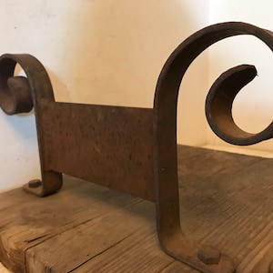 May include: Rustic metal shelf bracket with a decorative scroll design. The bracket is made of iron and has a dark brown patina. It is mounted on a wooden plank.