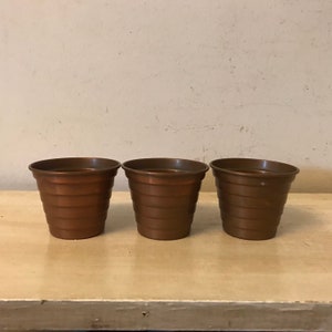 May include: Three small copper-colored pots with a ribbed design. The pots are arranged in a row on a light brown surface.