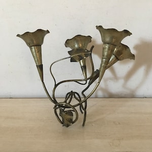 May include: A brass candlestick with a floral design. The candlestick has four candle holders shaped like trumpet flowers. The base of the candlestick is a stylized stem with leaves.