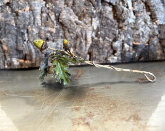 May include: A handcrafted metal sculpture of an oak branch with two acorns and green leaves. The branch is painted white with a twisted design and a loop at the end. The acorns have a yellow-green cap. The sculpture is set against a textured wood background.
