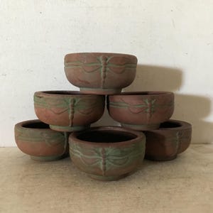 May include: Six small terracotta pots with a dragonfly design. The pots are stacked in a pyramid shape.