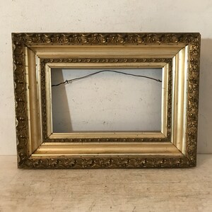 May include: A gold-colored picture frame with an ornate, detailed design. The frame has a rectangular shape and a slightly raised edge.