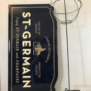 May include: A dark blue metal sign with gold lettering and trim, advertising "ST-GERMAIN" liqueur. The sign includes text "Enjoy ST-GERMAIN and CHAMPAGNE" and a bicycle illustration. A tall, metal, spherical sculpture stands beside the sign.