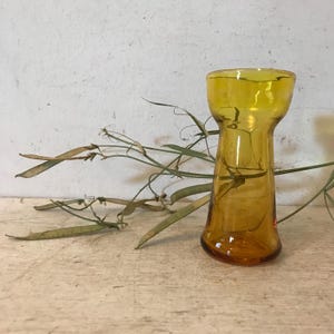 May include: A small, amber-colored glass vase with a narrow neck and a wide base. The vase is sitting on a light wood surface.