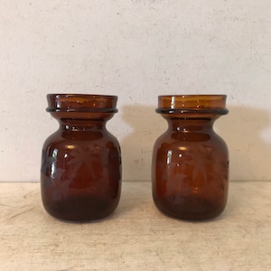 May include: Two amber glass bottles with etched designs. The bottles have a wide mouth and a narrow neck.