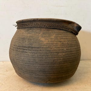 May include: A rustic, round, earthenware pot with a textured, ridged surface. A woven rope detail encircles the top rim. The pot is a muted brown color, suggesting an aged or antique appearance. The pot is likely used for storage or display.