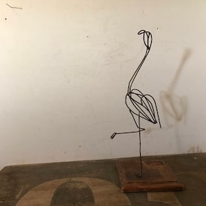 May include: A wire sculpture of a flamingo standing on a wooden base. The sculpture is made of thin wire and is painted black. The base is made of wood and is painted brown.