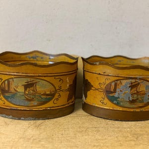 May include: Two antique, yellow metal containers with scalloped rims. Each container features an oval painting of sailboats on a body of water. The containers have a brown base and are decorated with a vine-like design.
