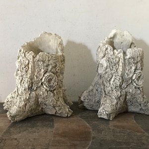 May include: Two off-white tree stump planters with textured bark-like surfaces. Each planter has a hollowed-out top and a decorative rose-like detail. The planters are suitable for indoor or outdoor use.