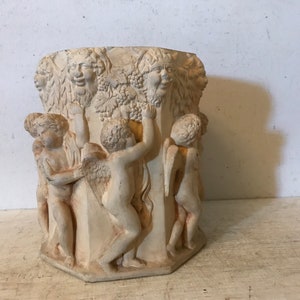 May include: A white ceramic planter with a hexagonal shape, featuring a relief design of cherubs and a vine with grapes. The planter has a rustic, antique look.