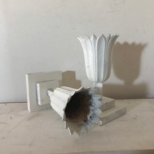 May include: Two white ceramic tulip-shaped candle holders. The holders are attached to square bases. The holders are in a tilted position.