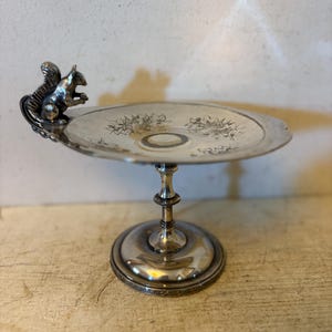 May include: Silver-toned pedestal dish with a decorative squirrel handle. The dish features floral etchings on the plate and a detailed squirrel figurine. The base is round and ornate, adding to its vintage charm.