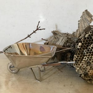 May include: A silver miniature wheelbarrow with a handle and a single wheel. The wheelbarrow is sitting on a wooden surface next to a piece of dried honeycomb.