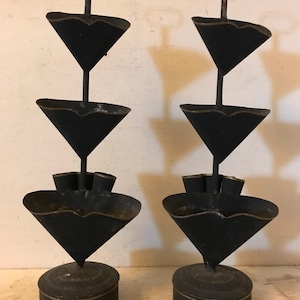 May include: Two antique black metal candle holders. Each has a round base, a conical midsection, and a flared top section. The top has a handle. The metal has a distressed finish, giving it a vintage appearance.