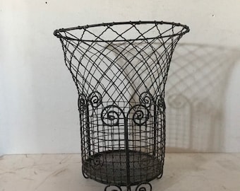 May include: A vintage-style, dark metal wire basket with a flared top and decorative scrollwork. The basket has a round base and is supported by curved feet. The wire mesh design allows for visibility of the contents. The basket is suitable for storage or display.