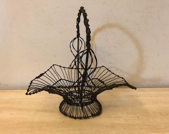 May include: A black wire basket with a twisted wire handle. The basket is empty and has a rounded bottom.