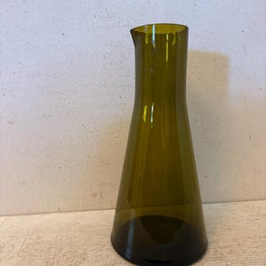 May include: A tall, olive-green glass vase with a flared base and a narrow neck. The vase has a slightly angled spout and is made of transparent glass, allowing light to pass through. The vase is likely used for displaying flowers or as a decorative item.
