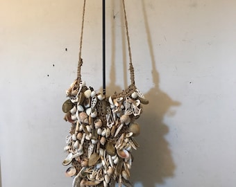 May include: A brown and white seashell bag with a long brown rope strap. The bag is filled with various seashells and is hanging from a black stand.