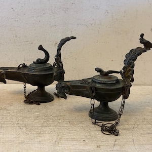 May include: Two antique bronze oil lamps with intricate detailing. Each lamp features a round base, a central bowl, and a spout. Decorative elements include curved handles, chains, and bird-like figures. The lamps are displayed on a light-colored wooden surface.