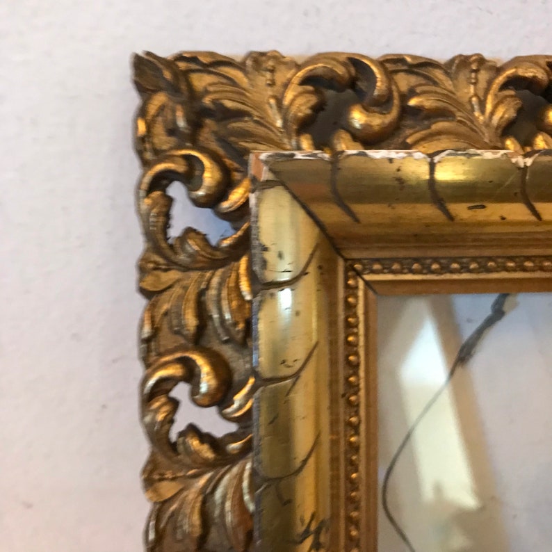K&ouml;nnte beinhalten: Ein verzierter Bilderrahmen in Gold mit einem detaillierten, geschnitzten Design. Der Rahmen hat eine leicht abgenutzte Optik, was seinen Vintage-Charme verst&auml;rkt.