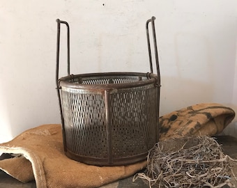 May include: A rusty metal wire basket with a round, open top and a woven wire design. The basket is sitting on a brown burlap surface.