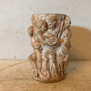 May include: A white ceramic vase with a relief sculpture of four figures. The figures are in a classical style and appear to be holding a fish or other animal. The vase is approximately 6 inches tall.