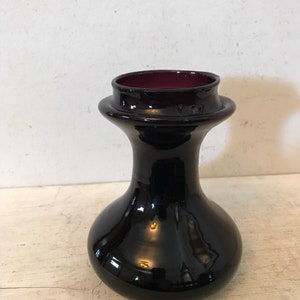 May include: A small, dark purple glass vase with a rounded body and a flared neck. The vase has a glossy finish.