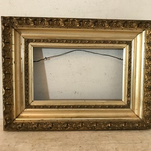 May include: A gold-colored ornate picture frame with a decorative border. The frame is made of wood and has a light brown finish.