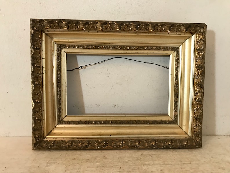 May include: A gold-colored ornate picture frame with a decorative border. The frame is made of wood and has a light brown finish.