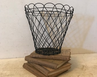 May include: A black wire basket with a scalloped top. The basket is sitting on a stack of books with the text "R. GUILLAUMIN-FLEURS JARDINS" on the top book.