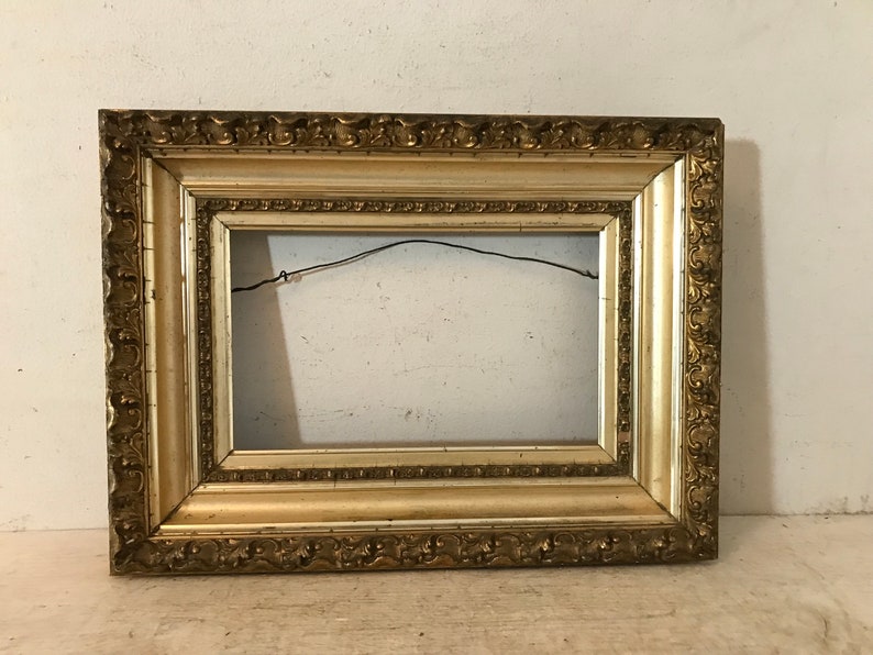 May include: A gold-colored ornate picture frame with a decorative border. The frame is empty and ready to display a picture or artwork.