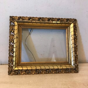 K&ouml;nnte beinhalten: Ein verzierter Bilderrahmen in Gold mit einem dekorativen, geschnitzten Design. Der Rahmen ist aus Holz und hat eine Glasfront.