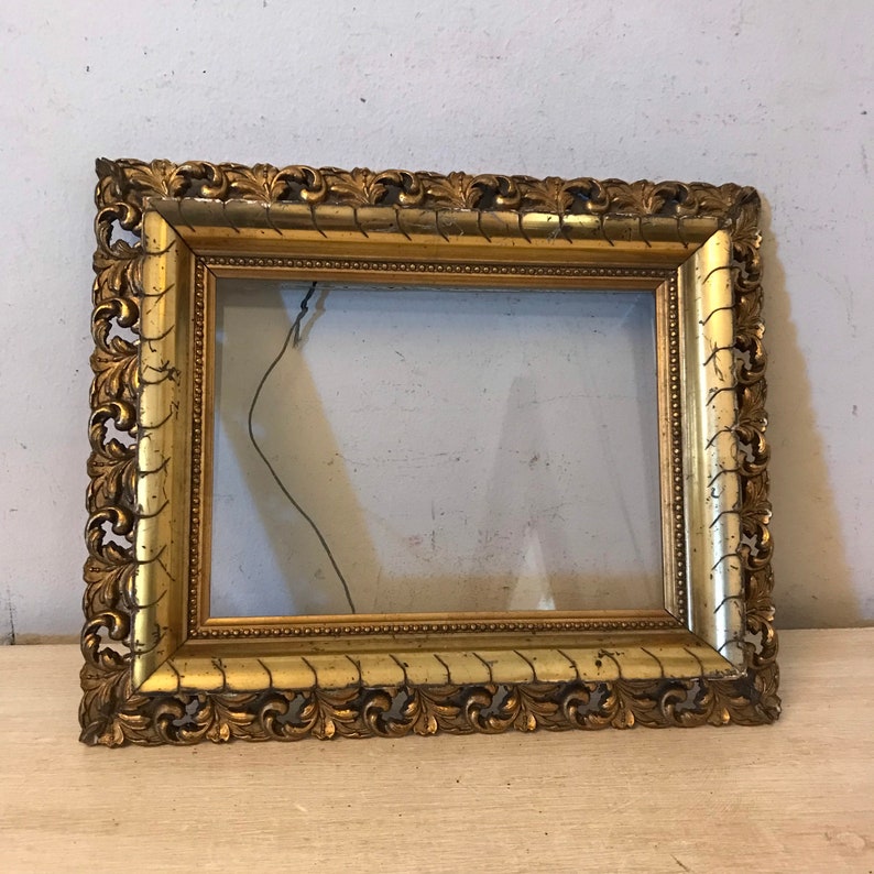 K&ouml;nnte beinhalten: Ein verzierter Bilderrahmen in Gold mit einem dekorativen, geschnitzten Design. Der Rahmen ist aus Holz und hat eine Glasfront.