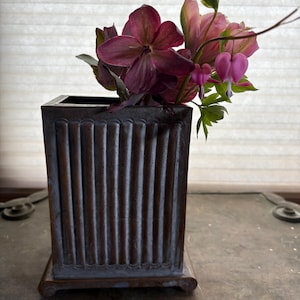 May include: A rectangular dark bronze-toned metal vase with vertical fluted detailing and a small base, containing a bouquet of deep purple hellebore flowers and pink bleeding heart stems with green foliage extending from the top opening.