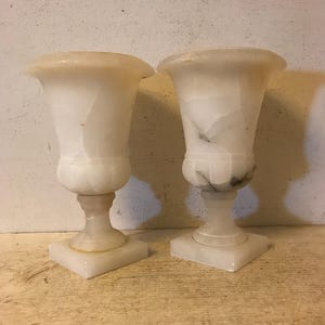 May include: A pair of white alabaster vases with a fluted design. The vases are on square bases and have a classic, elegant style.