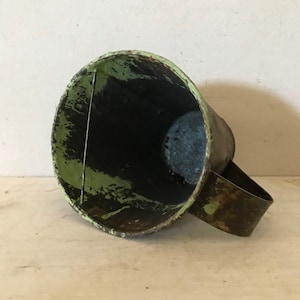 May include: A vintage metal cup with a green and black paint finish. The cup has a handle and is slightly dented.