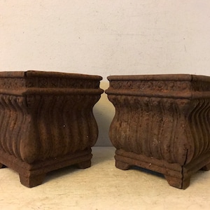 May include: Two antique, rusty, cast iron planter pots with a square top and a rounded bottom. The pots have a decorative, ribbed design.