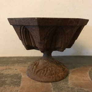 May include: A rustic, cast iron planter with a hexagonal top and a decorative base. The planter has a distressed, aged look and is perfect for adding a touch of vintage charm to your home decor.